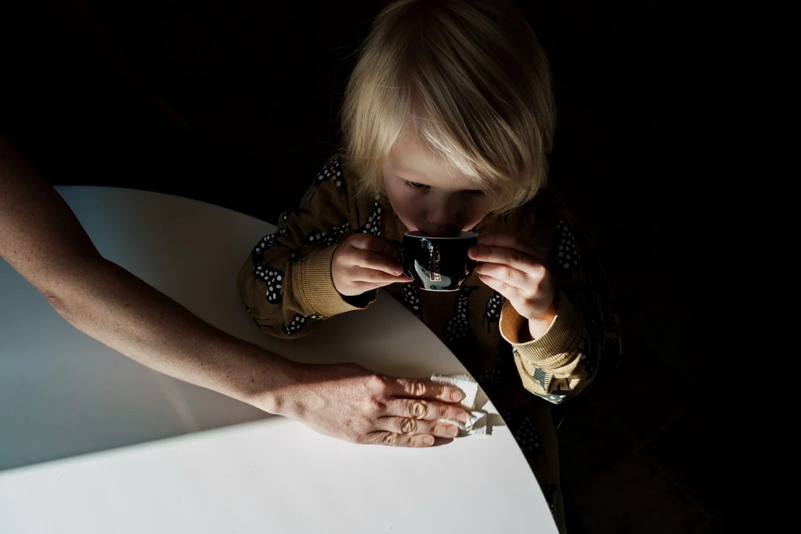 A child drinks from a small dark cup at a white table beside an adult’s arm.