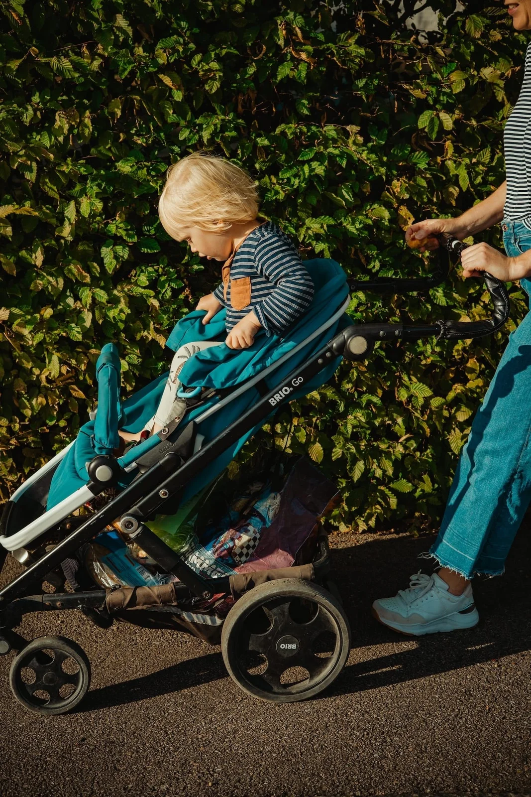 A child in a pushchair passes sunlit green foliage, with an adult partly visible beside them.