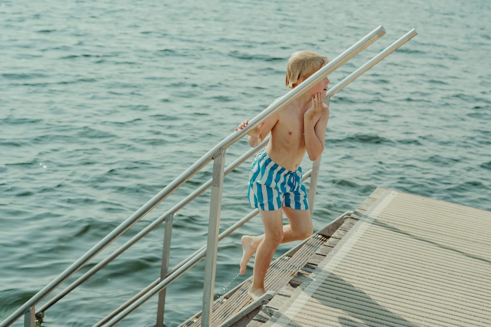 A child in blue striped swimming shorts climbs metal steps out of the water.