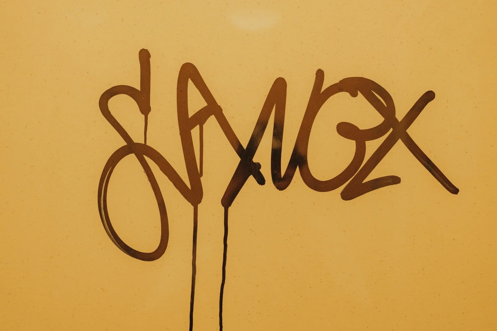Dark looping graffiti with drips of paint on a mustard-yellow wall.